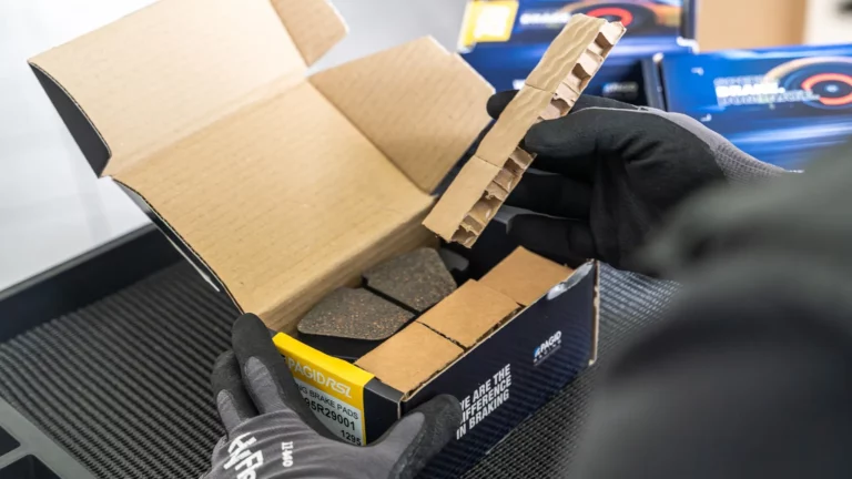Gloved worker placing a PAGID Racing motorsport brake pad set into a recycled cardboard box lined with honeycomb cardboard filler in place of polyurethane foam.